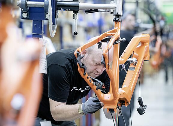 Ein Mitarbeitender montiert den orangefarbenen Rahmen eines Elektrofahrrads in einer Fahrradmanufaktur.