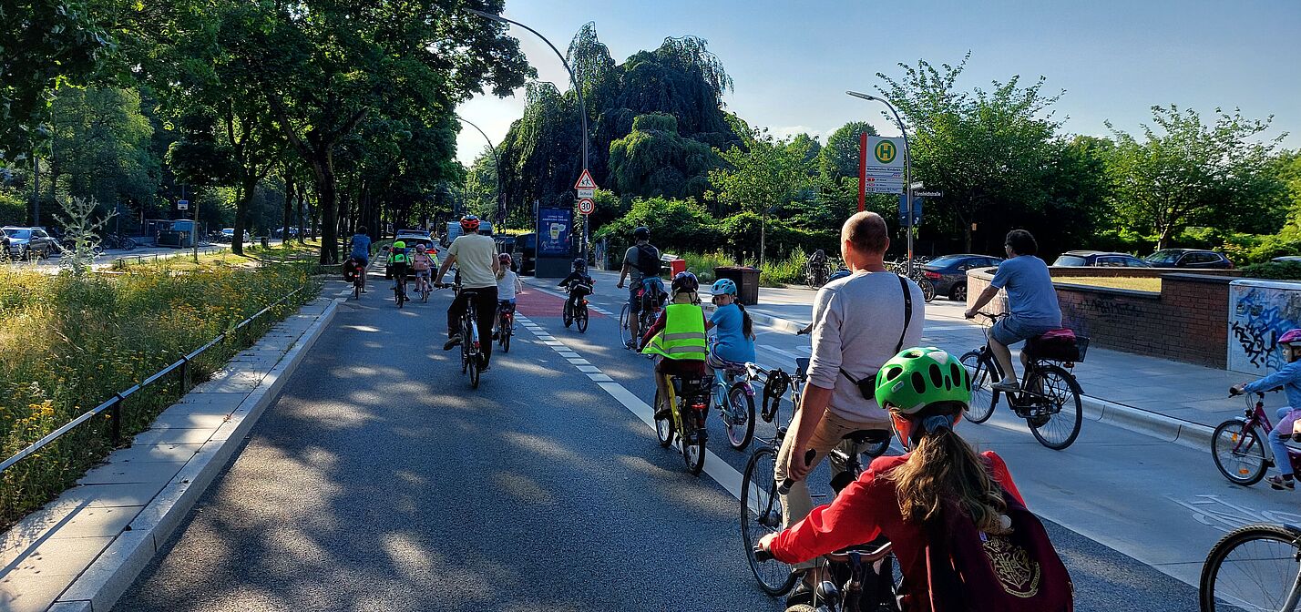 Eine Gruppe von Kindern fährt auf ihren Rädern auf der Straße. Sie werden von Erwachsenen begleitet.